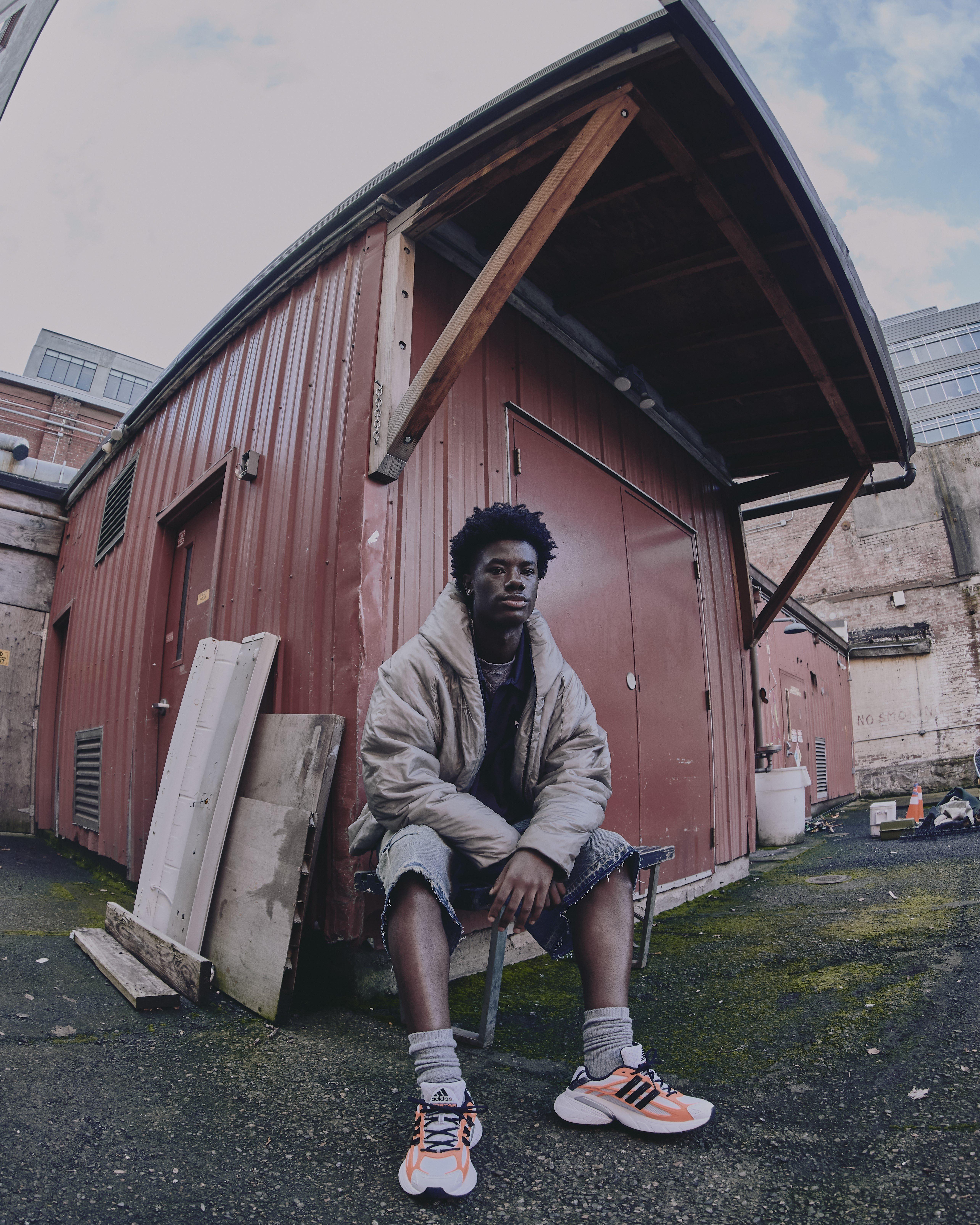 A low-angle, wide-shot of a man sitting on a bench outside a red corrugated metal building. He is wearing an oversized beige puffer jacket, denim shorts, grey socks, and orange/white Adidas Adistar XLG sneakers. The ground is damp asphalt with patches of moss, and the sky is visible under a wooden roof overhang.