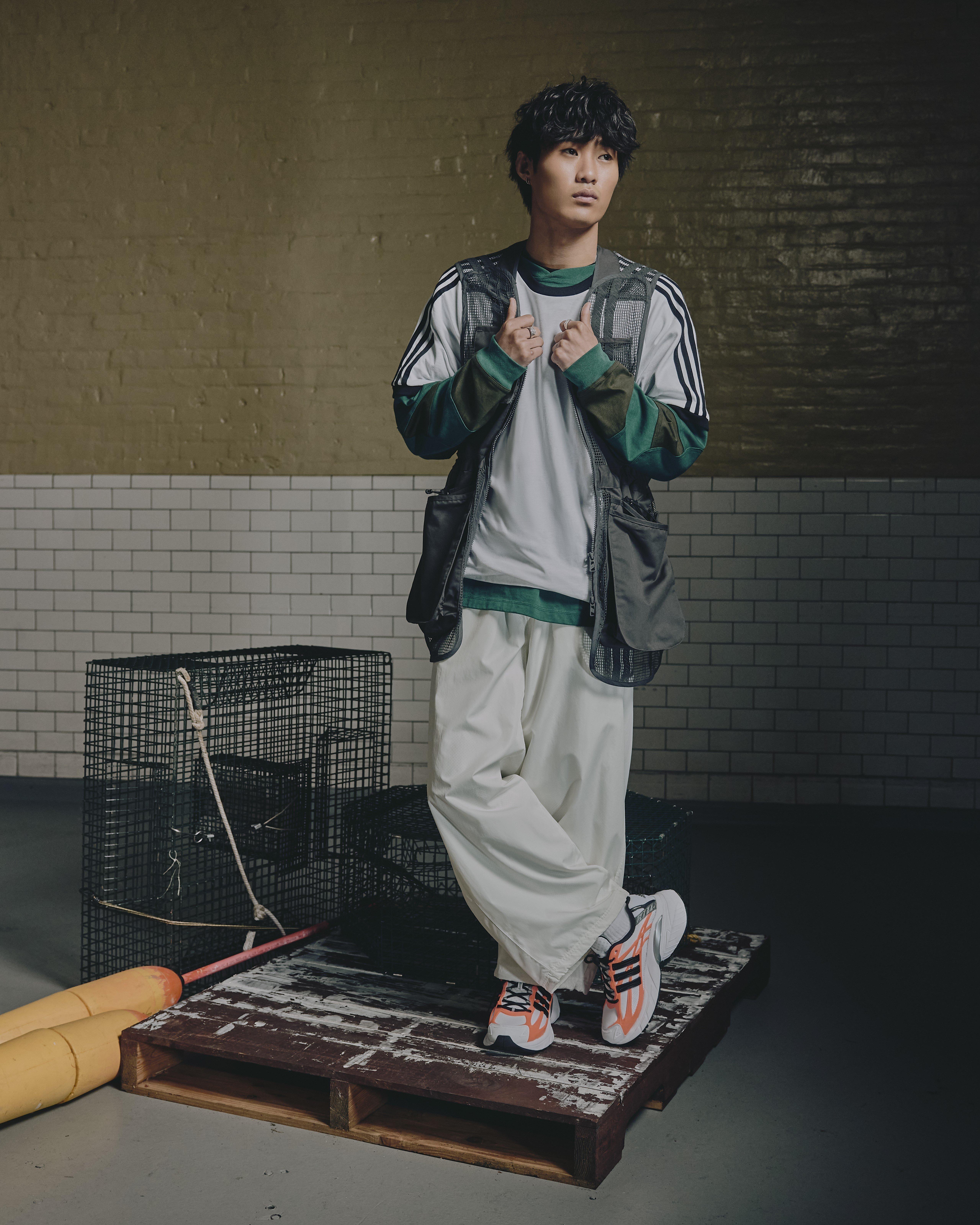 A full-body lifestyle shot of a man standing on a weathered wooden pallet in an industrial room with white tiled walls. He is wearing baggy white trousers, a white t-shirt, and a green-and-grey utility vest. On his feet are the orange and white Adidas Adistar XLG sneakers. Metal wire cages and yellow buoys sit on the floor beside him.