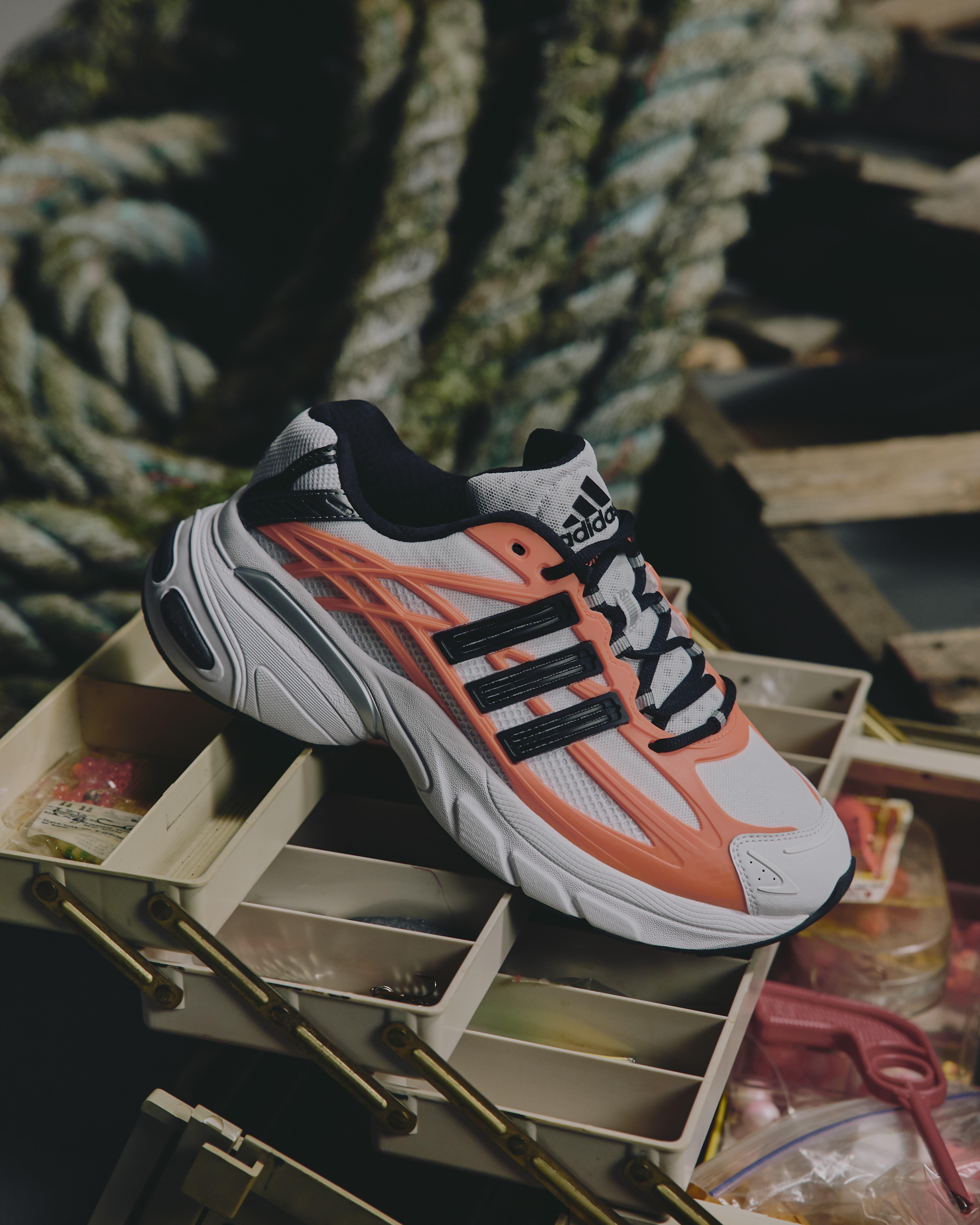 A side-profile product shot of an Adidas Adistar XLG sneaker. The shoe features a white mesh base with vibrant orange "cage" overlays, black three-stripes, and a chunky white midsole with silver accents. It is resting on a beige multi-compartment tackle box with thick, coiled green ropes in the blurred background.