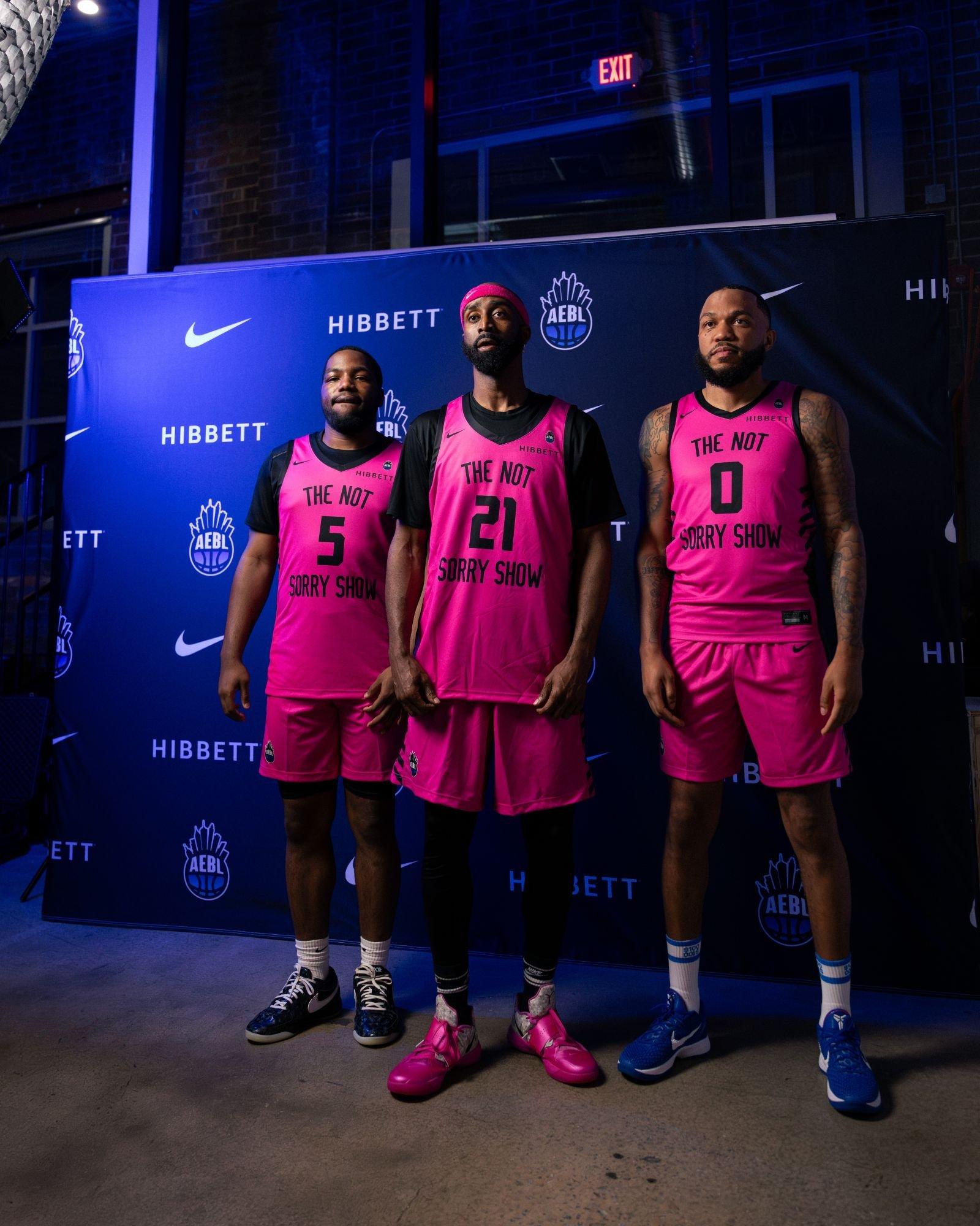 Three basketball players in bright pink uniforms pose in front of a Hibbett, Nike and AEBL step-and-repeat backdrop.