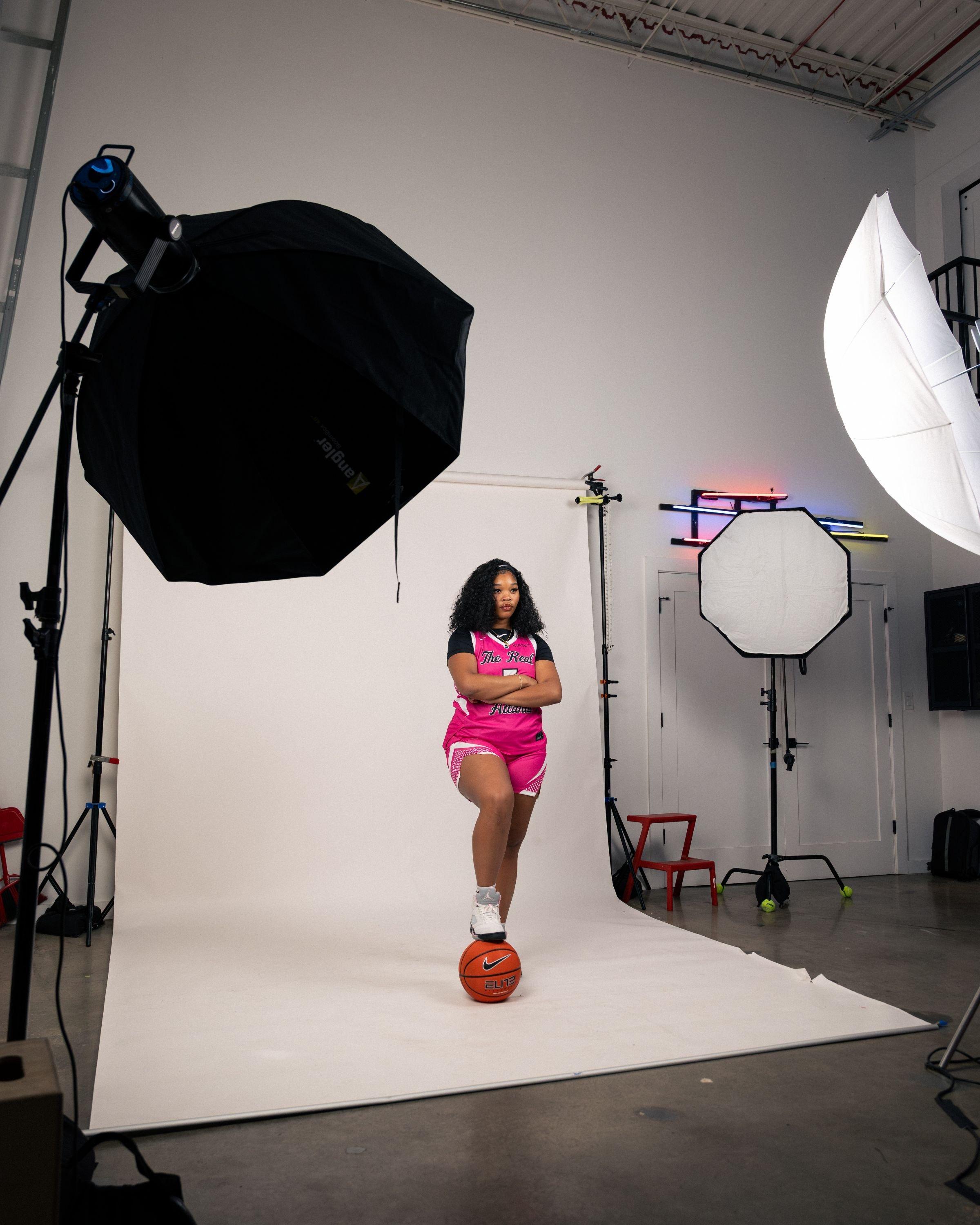 Basketball player in a pink uniform poses with one foot on a basketball during a studio photoshoot.