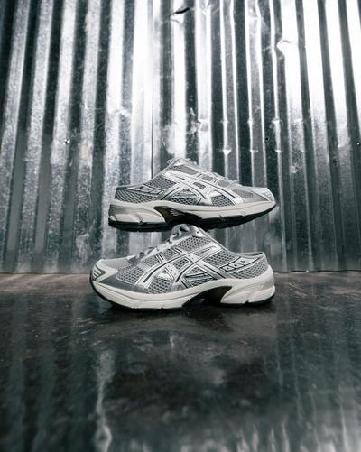 Pair of silver ASICS mesh sneakers stacked against a corrugated metal wall, showing side views with white midsoles and black outsole accents.