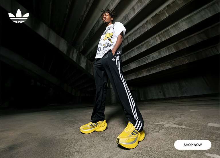 A promotional banner for Adidas Originals featuring a model in a streetwear outfit. The look includes a white graphic T-shirt with a distressed Adidas Originals logo and an angry yellow duck illustration, black track pants with white stripes, and bright yellow chunky sneakers with silver accents. The background is a dark, industrial concrete setting.