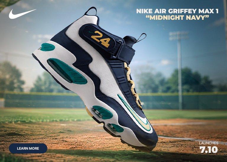 Nike Air Griffey Max 1 “Midnight Navy” sneaker displayed on a baseball field with batting cage netting and stadium lights in the background.