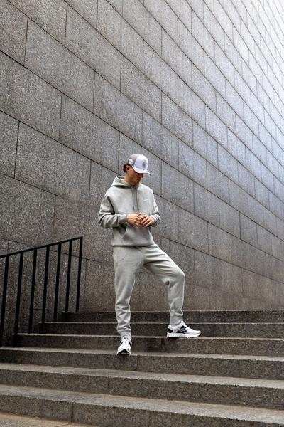 Man wearing a light gray hoodie and matching joggers with white sneakers, standing on concrete steps against a modern stone wall.