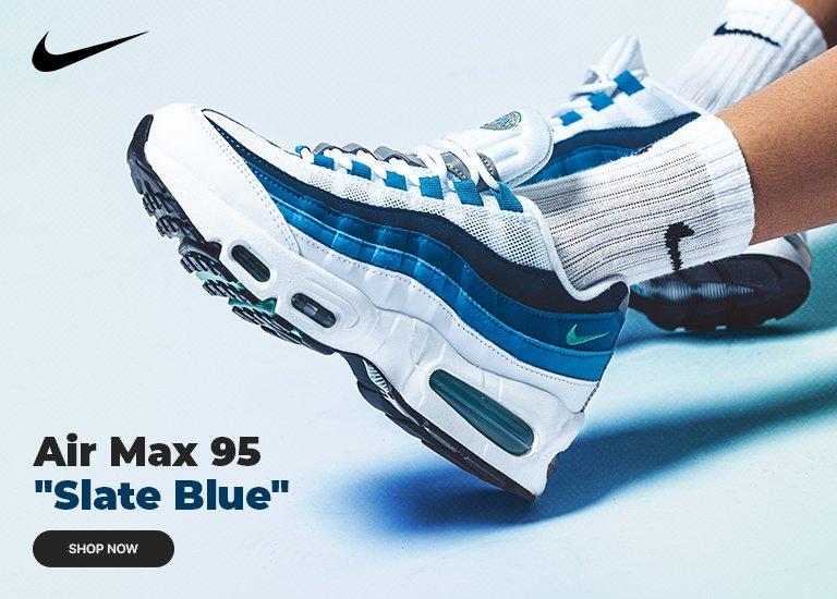 A promotional banner for Nike Air Max 95 Slate Blue sneakers. The image shows a close-up of the shoes being worn with white Nike socks against a light blue background. A Nike swoosh is in the top left corner, with the product name and a black shop now button in the bottom left.