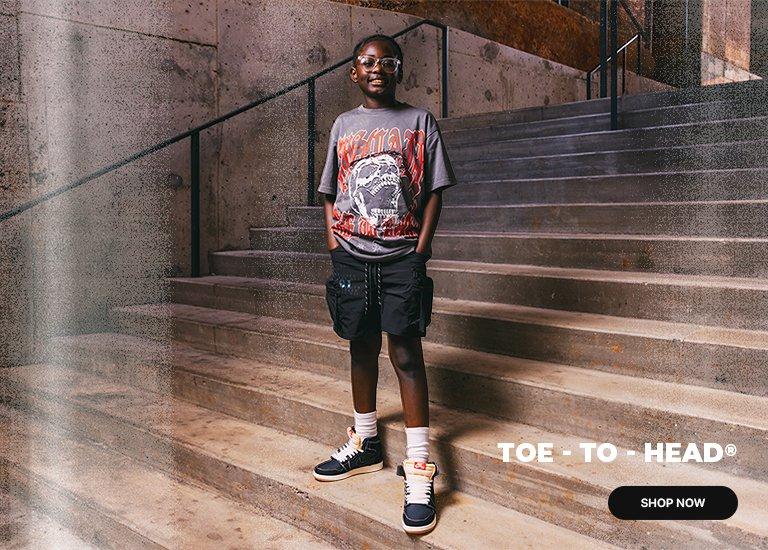 A split-screen promotional image featuring a young person wearing glasses, a graphic t-shirt, shorts, and high-top sneakers. On the left, they stand on concrete stairs, and on the right, they are seated with a smile. The center text reads TOE - TO - HEAD repeated three times, and a SHOP NOW button is located in the bottom right corner.