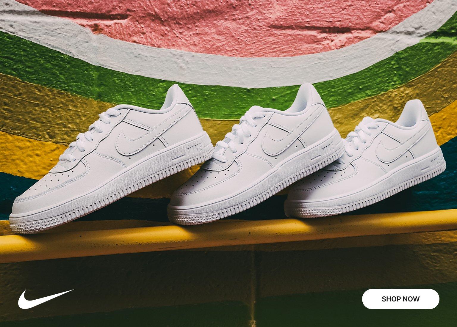 Three pairs of all-white Nike Air Force 1 sneakers lined up by size from largest to smallest against a brightly painted wall. Features a white Nike swoosh and a Shop Now button.