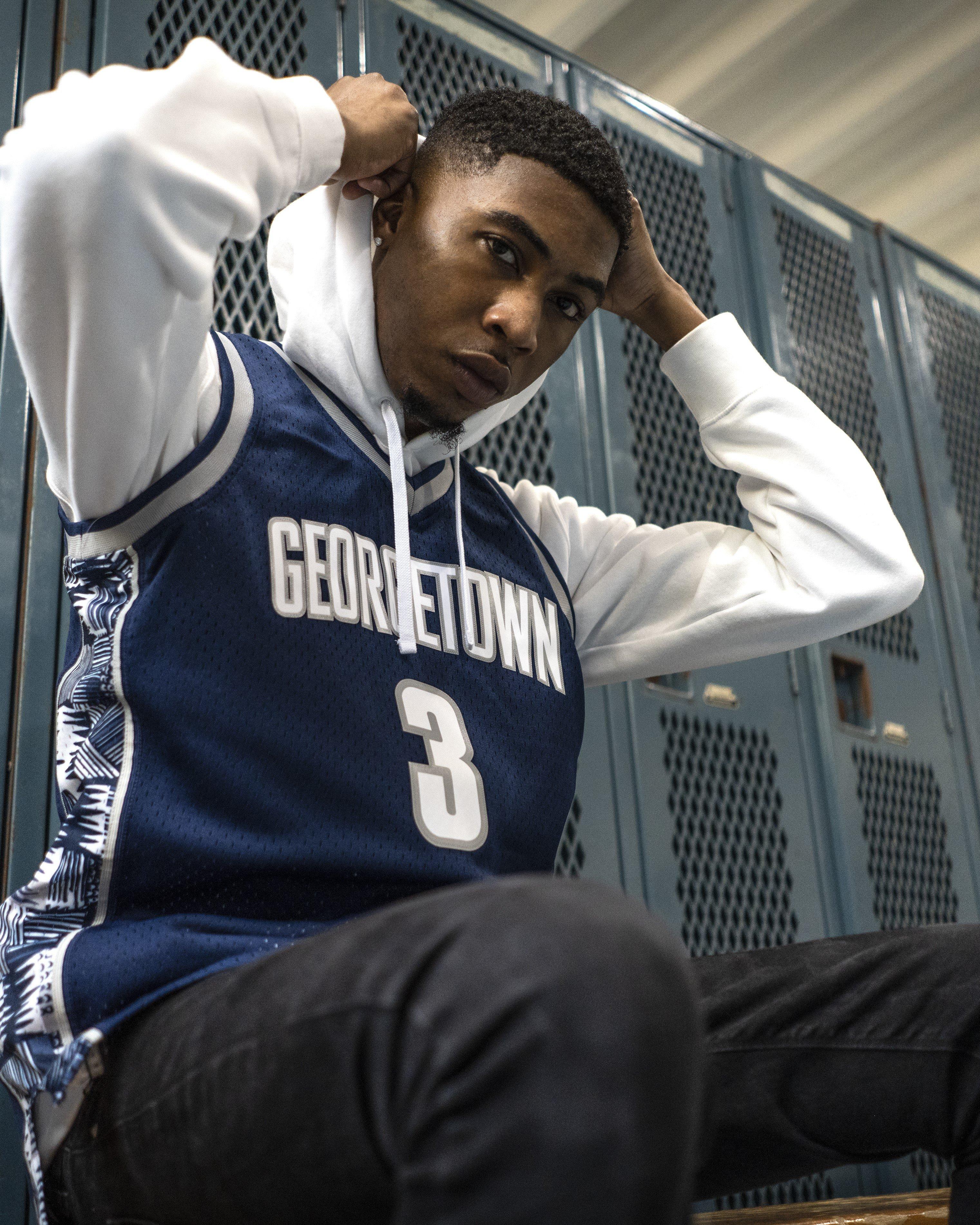 A young Black man sits on a wooden bench in a locker room, wearing a blue Georgetown University basketball jersey with the number 3 over a white hoodie. He is looking directly at the camera while holding the sides of his hood. The background consists of rows of blue metal lockers.