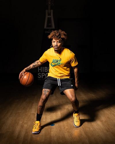 A man with curly hair wearing a gold t-shirt and black shorts dribbles a basketball on a wooden court. He is also wearing gold and black basketball sneakers. The background is dark, with a "Spalding" sign and a "Hibbett Sports" stand visible.