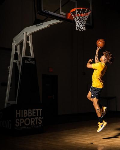 A man in a gold shirt and black shorts jumps toward a basketball hoop, balancing a basketball on his finger. The background is a dimly lit gym with a basketball hoop and a "Hibbett Sports" stand.