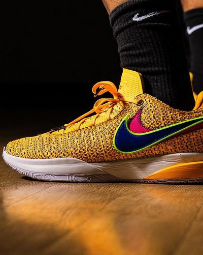 A close-up side view of a Nike LeBron 20 shoe. The shoe is a golden knit material with a multi-colored Swoosh: pink with a blue outline, and a lime green trim. The laces are orange, and the sole is white, gray, and yellow. The shoe is on a wooden basketball court.