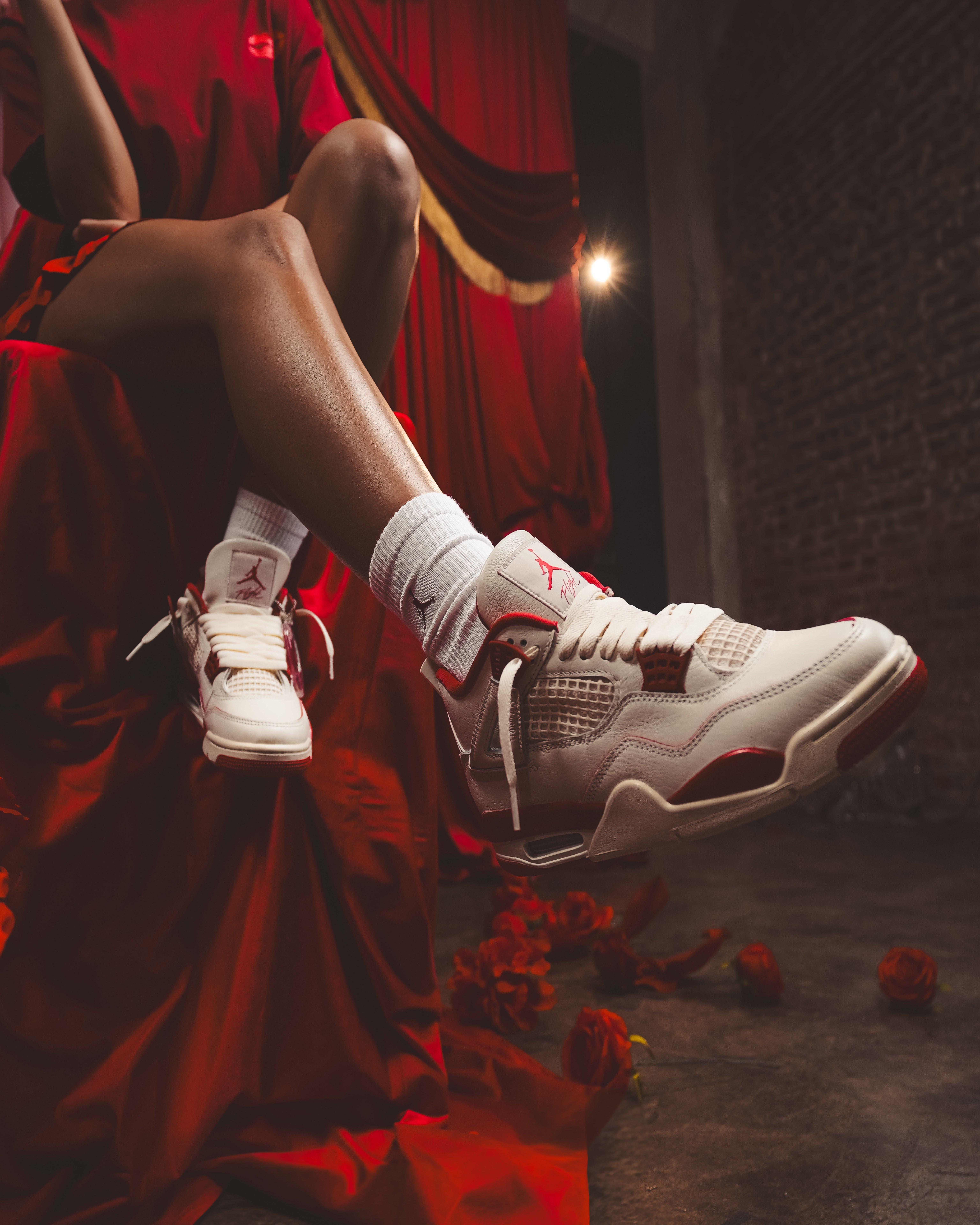 A low-angle, focused shot of the woman&rsquo;s legs and feet. She is wearing white ribbed socks and the Air Jordan 4 sneakers. One foot is extended toward the camera, highlighting the intricate mesh and leather details of the shoe. She is seated on a red draped fabric in a dimly lit room with rose petals on the floor.
