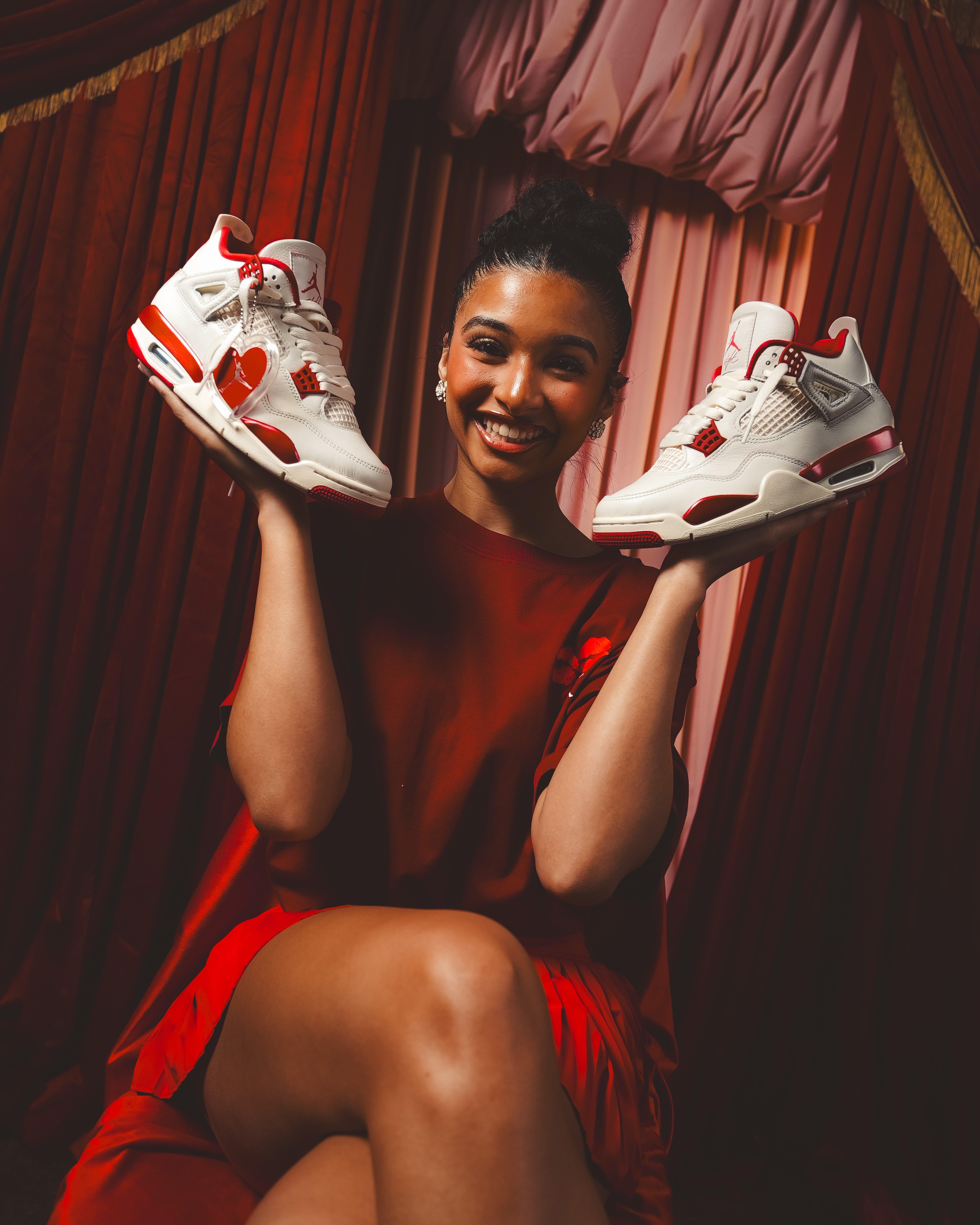 A smiling woman with her hair in a high bun sits against a backdrop of deep red and pink theatrical curtains. She is holding up a pair of Air Jordan 4 "Valentine's Day" sneakers, one in each hand, showcasing their white leather and metallic red accents.