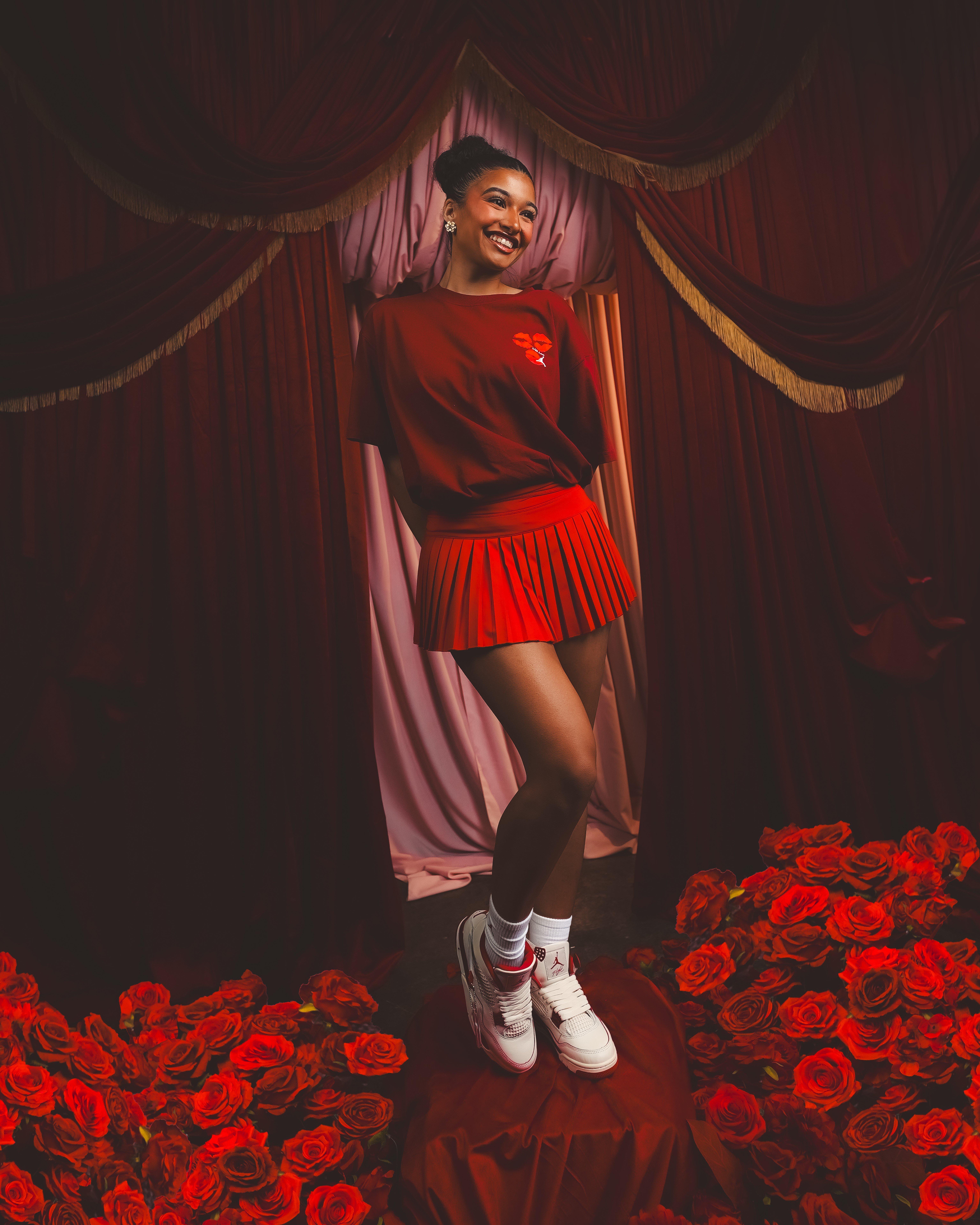 A full-length shot of the woman standing on a platform surrounded by a sea of red roses. She is wearing a red t-shirt, a pleated red mini skirt, white crew socks, and the white and red Jordan 4 sneakers. The background features elegant, draped red curtains.