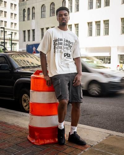 Model wearing a cream Palm Angels graphic T-shirt, gray cargo shorts, white Nike socks and black sneakers on a city sidewalk.