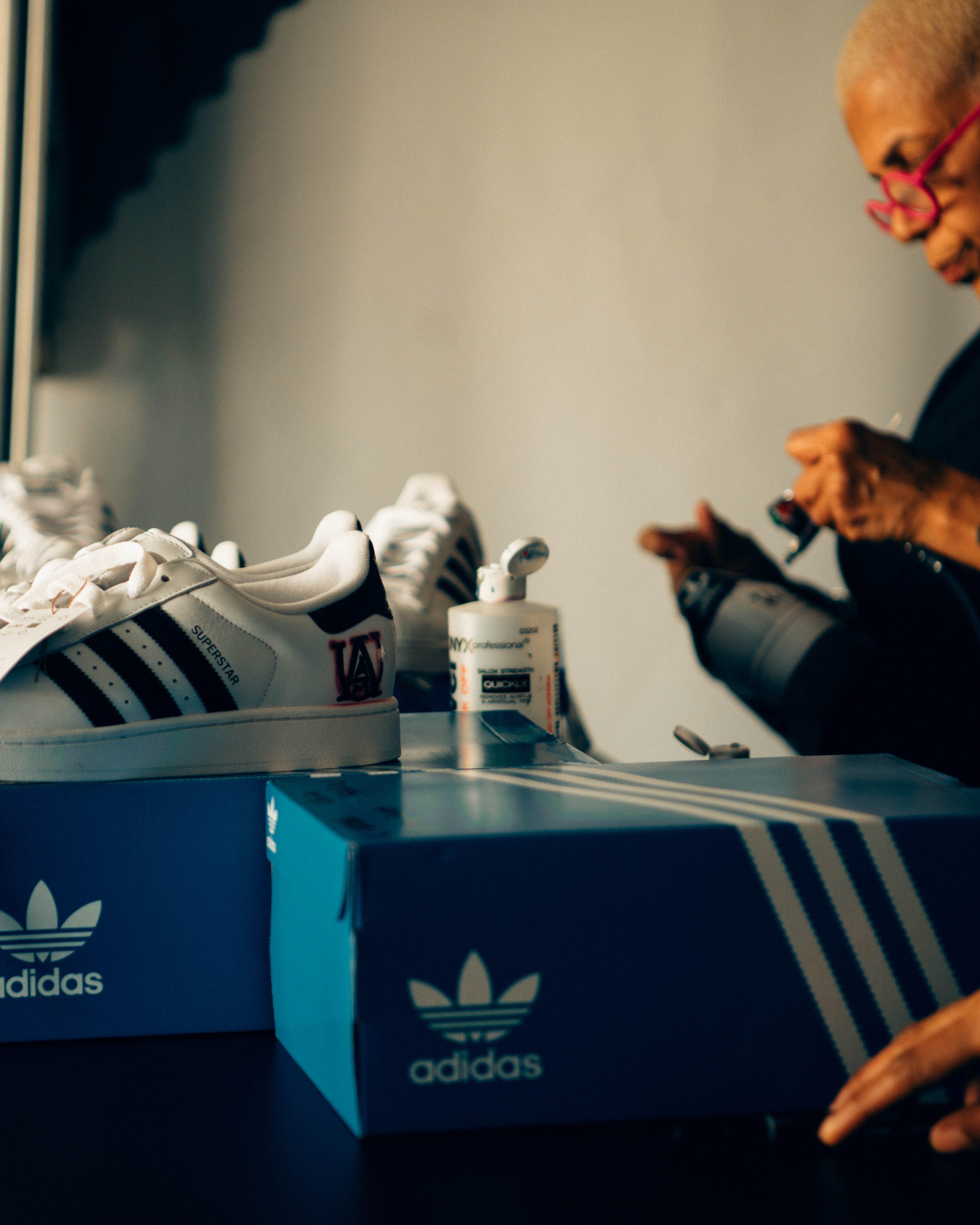 A A display of custom and classic adidas Superstar sneakers on a counter inside the Hibbett tent, including a pair in red and white and a pair being customized. boy wearing glasses, an Alabama football jersey, and face paint that reads "BAMA" is cheering with his mouth open. He is wearing a lanyard that says HIBBETT and BALLIN IN BOUTWELL.