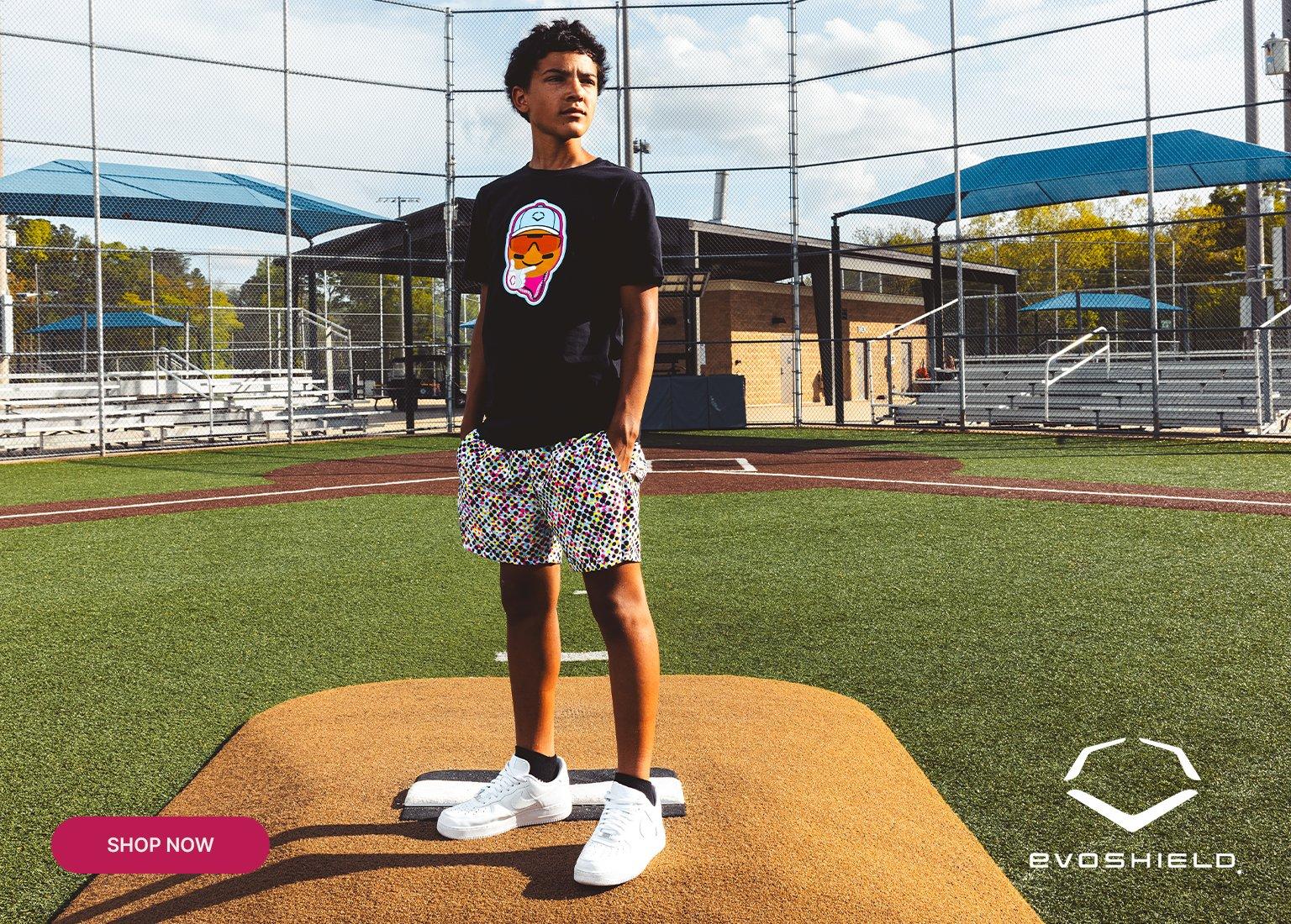 A young baseball player stands on a pitcher's mound wearing a black EvoShield graphic tee with a colorful face emblem, patterned multi-colored shorts, and white sneakers. The background features a sunny baseball field with bleachers and a chain-link fence. An EvoShield logo is in the bottom right corner, and a magenta "SHOP NOW" button is in the bottom left.