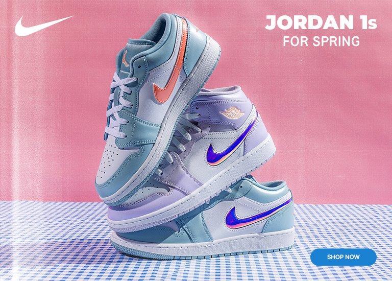 A stack of three Jordan 1 sneakers in pastel shades of blue, purple, and orange. The shoes sit on a blue-and-white checkered surface against a pink backdrop with a "Jordan 1s FOR SPRING" headline. A Nike swoosh is in the top left corner, and a blue "SHOP NOW" button is in the bottom right.