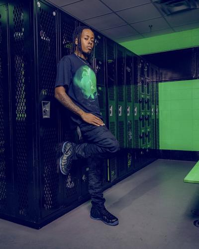 A young man with dreadlocks leans against a row of black metal lockers in a room with green walls, wearing a black graphic t-shirt and black Nike Air Foamposite sneakers.