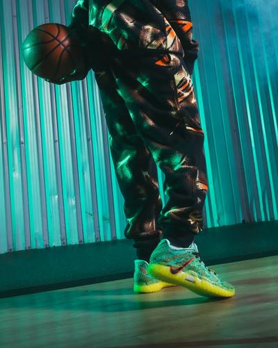 A person wearing a patterned black and orange tracksuit holds a basketball while standing on a court. They are wearing bright green and yellow Nike Cosmic Unity 3 sneakers, illuminated by teal studio lighting against a corrugated metal backdrop.