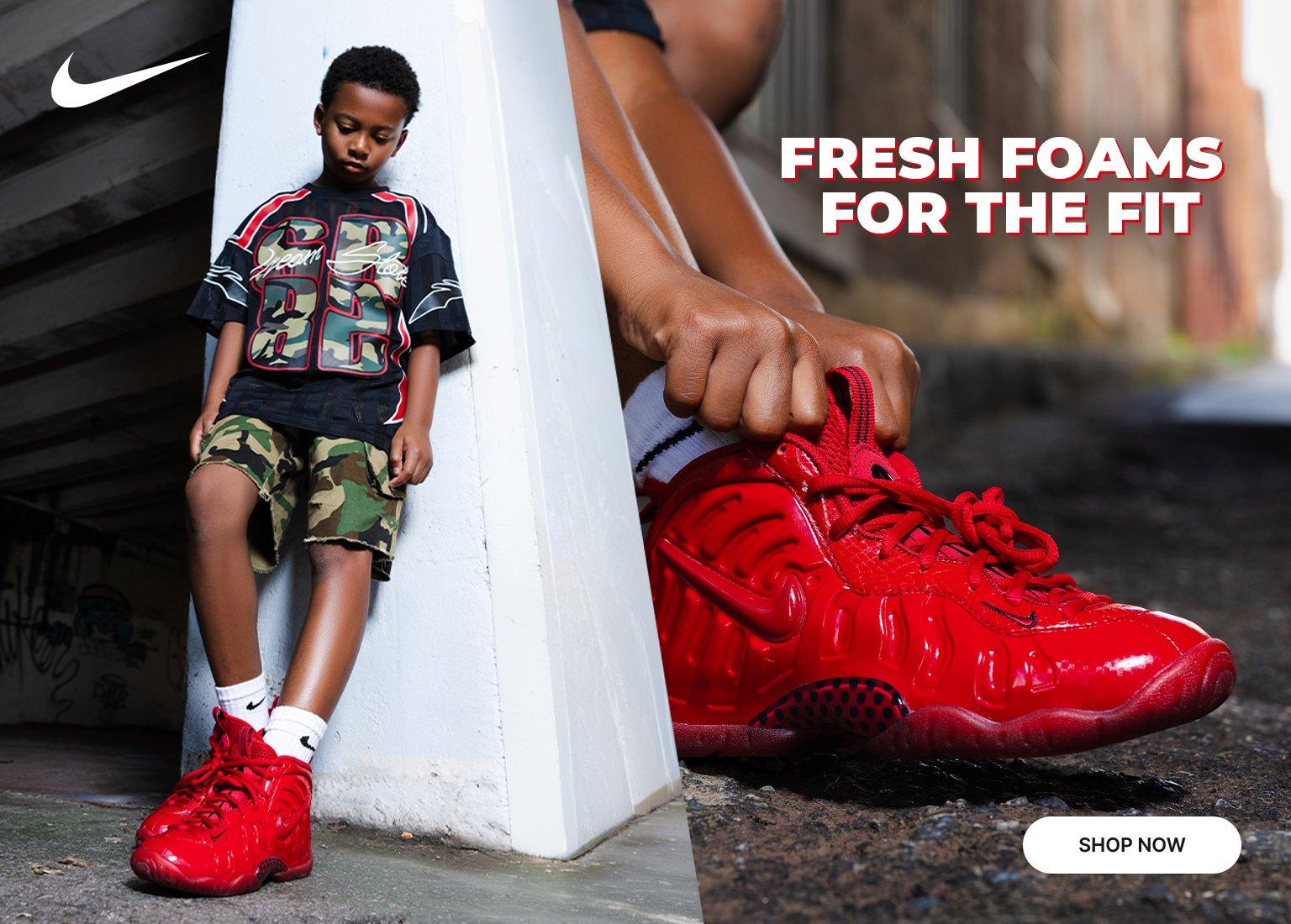 Alt text: A split-screen Nike promotional banner. On the left, a young boy leans against a concrete pillar wearing a black and camo graphic shirt, camo shorts, white socks, and red Nike Air Foamposite Pro sneakers. On the right, a close-up shows a hand adjusting the collar of the bright red Foamposite sneaker. Text reads "Fresh Foams For The Fit" with a "Shop Now" button below.