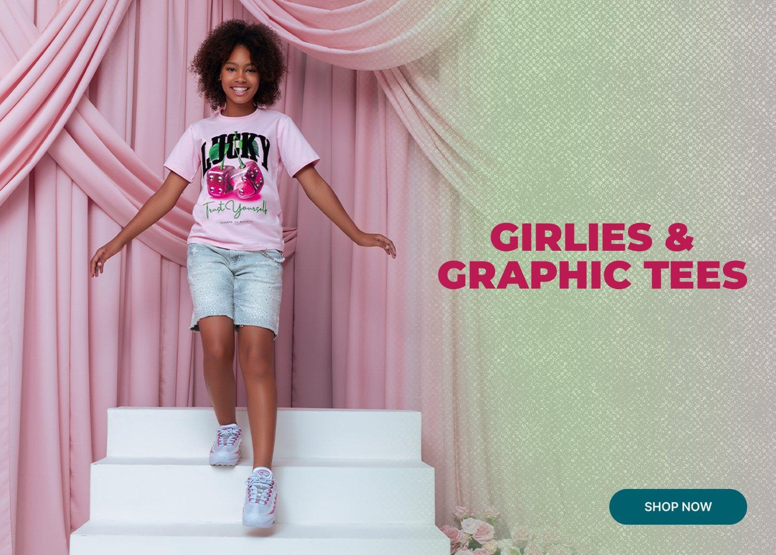 She is wearing a light pink graphic t-shirt featuring the word "LUCKY" and dice, paired with light-wash denim shorts and white sneakers. The top left corner displays the text GIRLIES & GRAPHIC TEES in maroon, and a teal SHOP NOW button is located in the bottom right.