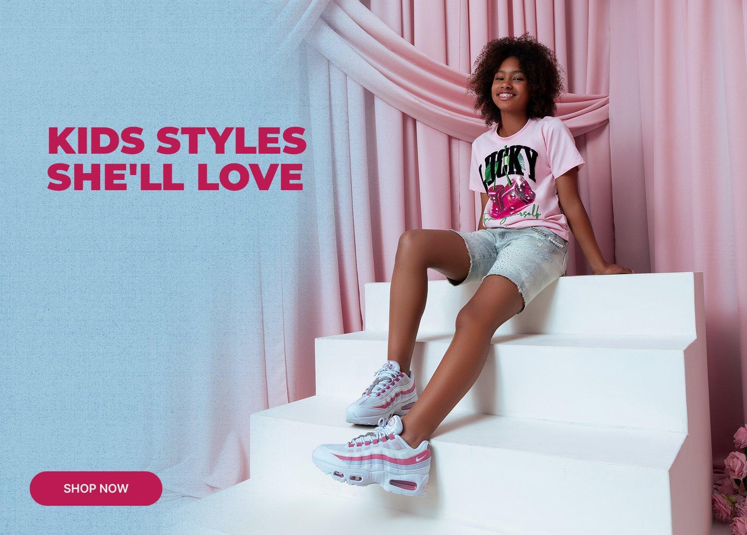 A Nike promotional banner titled KIDS STYLES SHE'LL LOVE featuring a young girl posing on white steps against pink draped curtains. She is wearing a pink graphic T-shirt with dice and cherries, denim shorts, and pink and white Nike sneakers. A Shop Now button is in the corner.