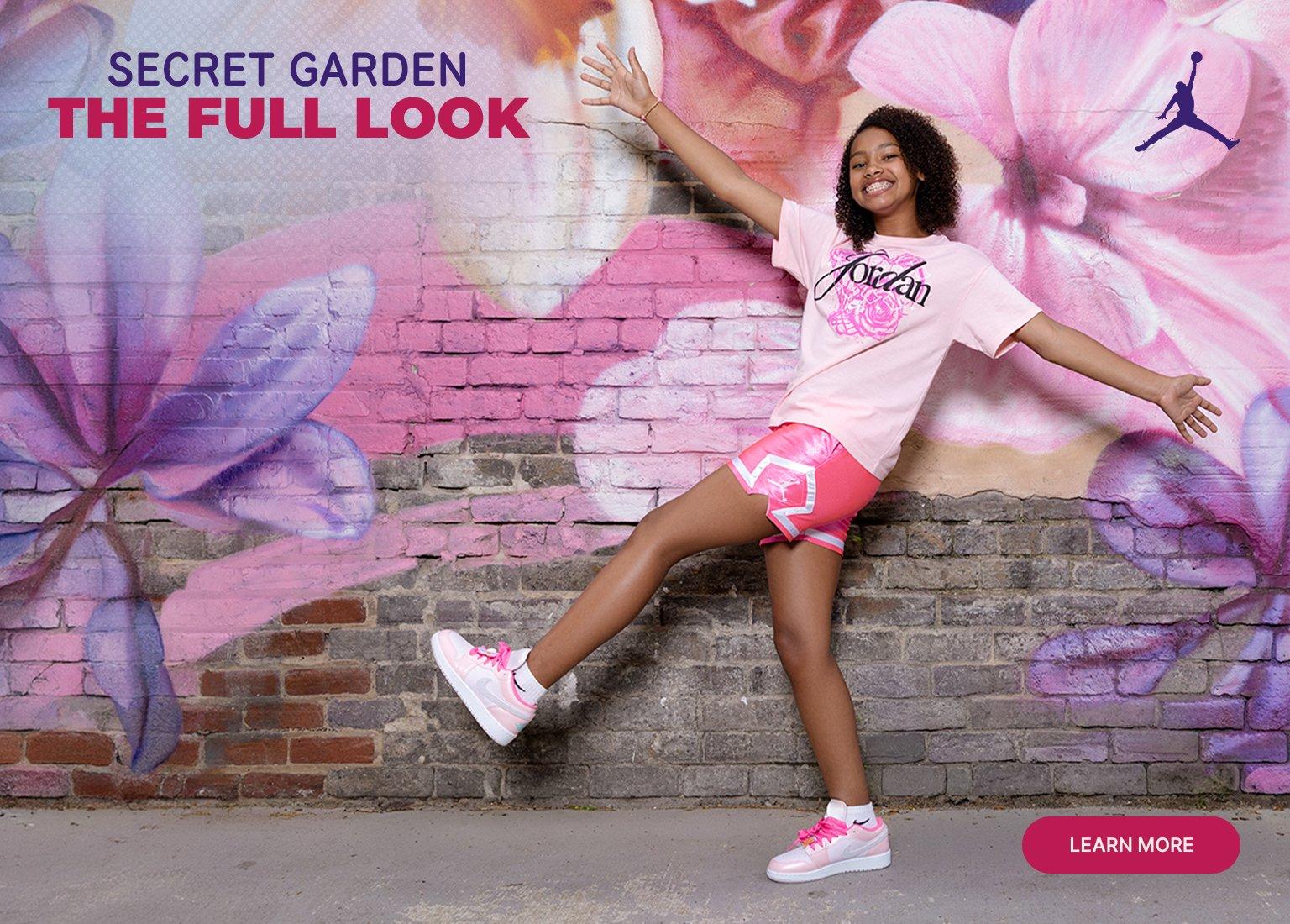 A young girl smiling and posing in front of a floral mural, wearing a pink Jordan "Secret Garden" graphic t-shirt, shiny pink athletic shorts, and pink and white Jordan sneakers. The image includes the text "SECRET GARDEN THE FULL LOOK," a purple Jumpman logo, and a "LEARN MORE" button.