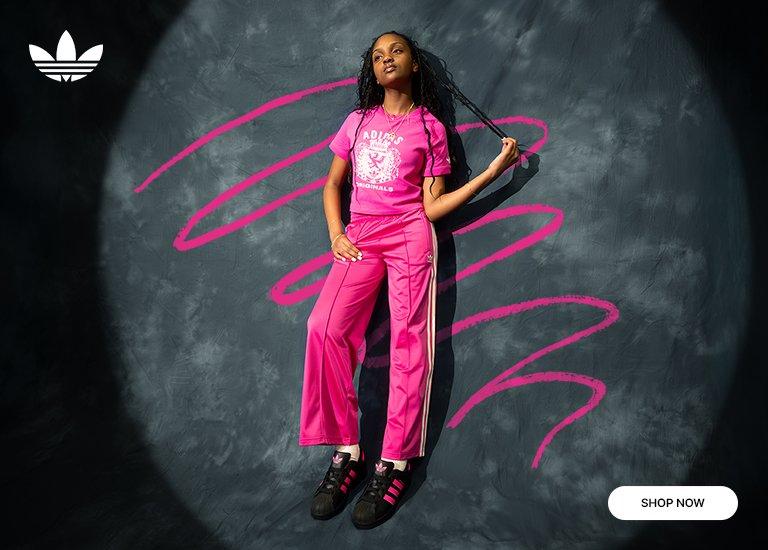 Young woman wearing a bright pink Adidas Originals graphic t-shirt and matching pink track pants with white side stripes, paired with black Adidas sneakers featuring pink accents. She poses against a dark textured backdrop with bold pink brushstroke graphics and the Adidas Originals logo in the top left corner, with a “Shop Now” button in the bottom right.