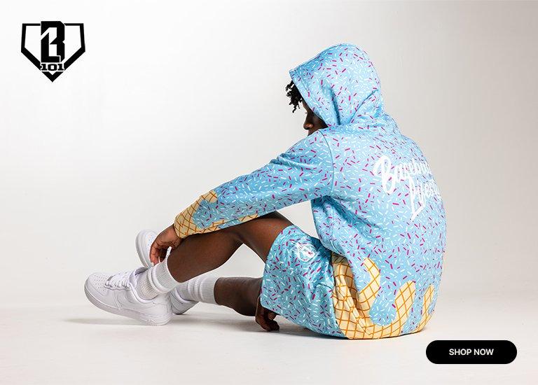 This promotional graphic for Baseball Lifestyle 101 features a person seated on a white floor, facing away from the camera to showcase an ice cream-themed apparel set. The light blue hoodie and matching shorts are covered in a colorful sprinkle pattern with waffle cone graphics at the hem and sleeves. The person is also wearing white socks and white sneakers. A "SHOP NOW" button is located in the bottom right corner, and the brand's home plate-shaped logo is in the top left.