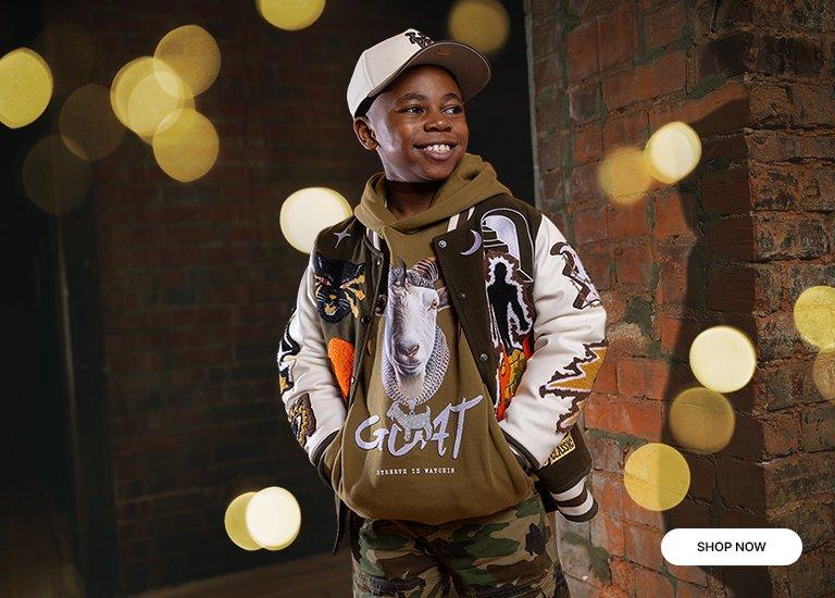 A young boy models a green and white varsity jacket with elaborate patches, including a large tiger on the back, and a matching baseball cap, standing against a brick wall.
