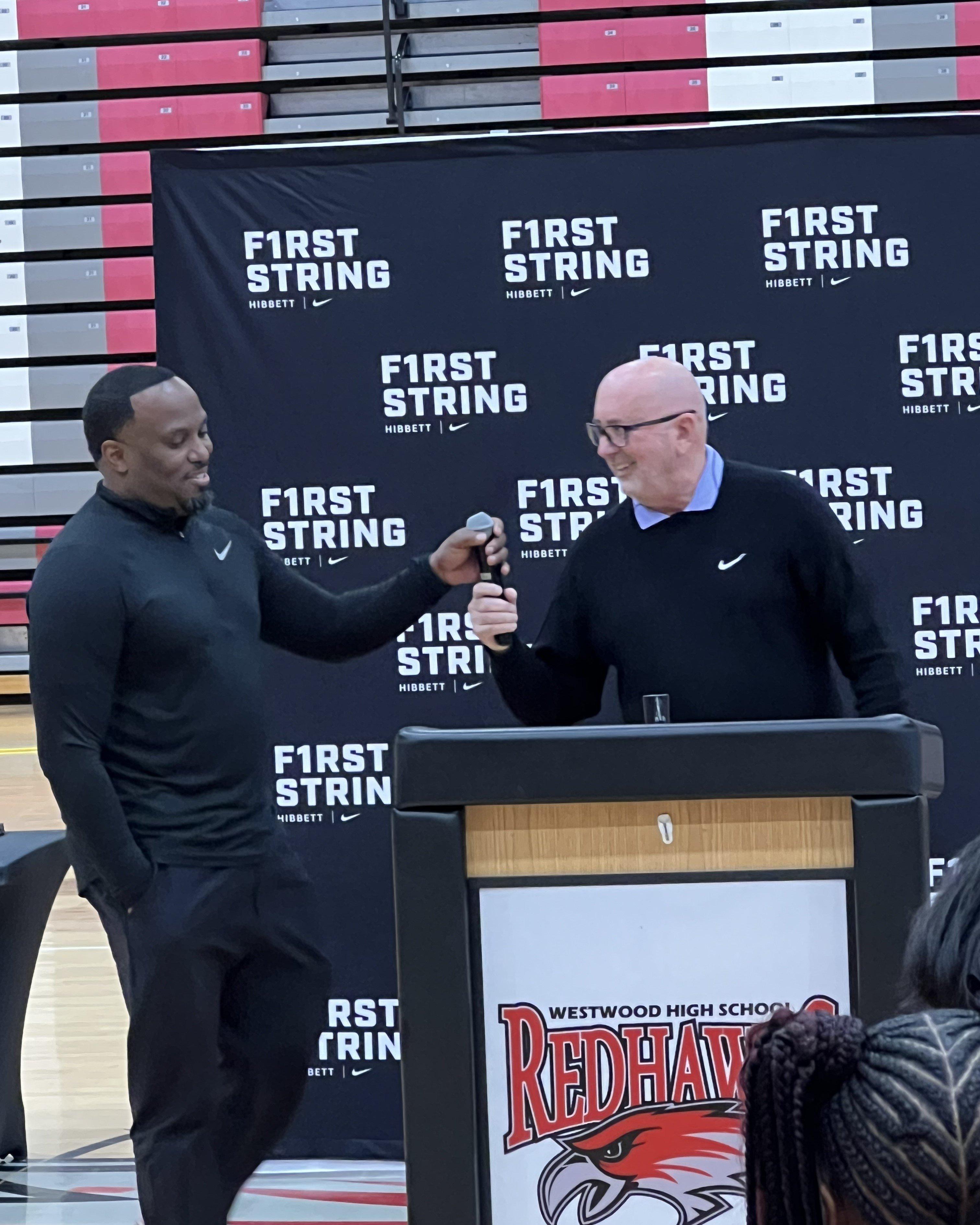 Two men speaking at a podium featuring the Westwood High School Redhawks logo, with a Hibbett and Nike First String backdrop behind them.