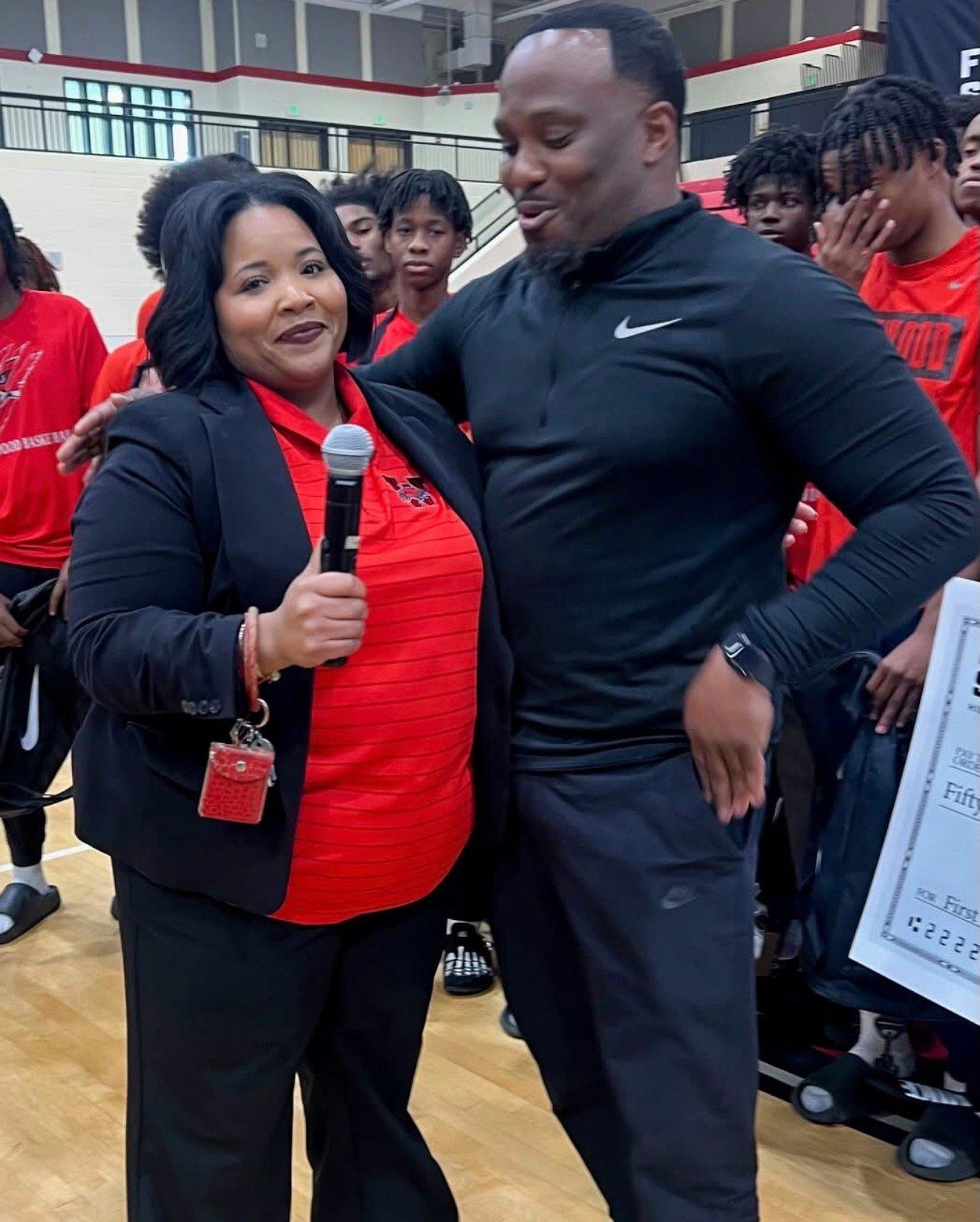 woman holding a microphone stands alongside a man in athletic wear, smiling and addressing the student-athletes during the First String presentation.