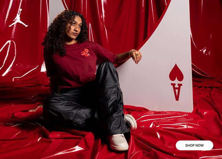 A woman with curly hair sits on a draped red backdrop next to a giant Ace of Hearts playing card. She is wearing a maroon Jordan t-shirt with a lipstick kiss graphic and black cargo pants.