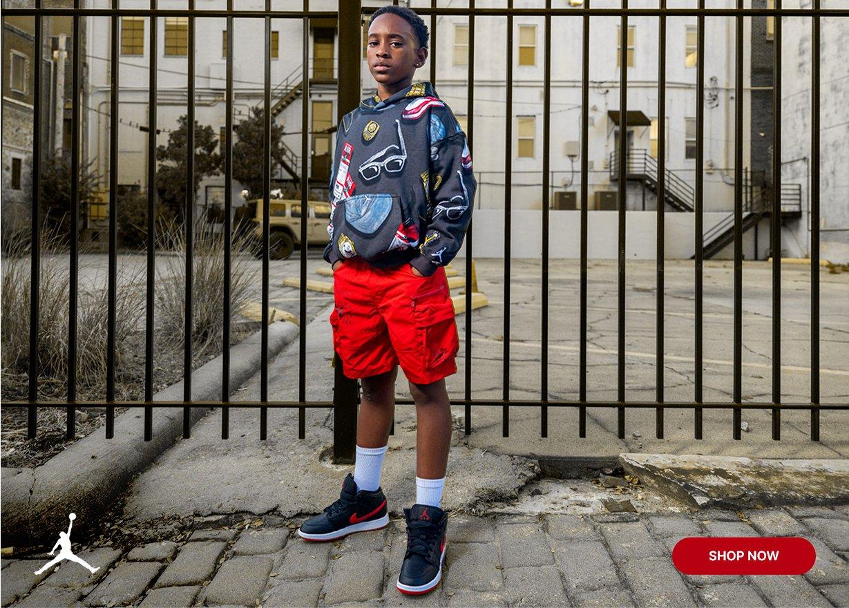 Young athlete wearing a Jordan graphic hoodie, red shorts and Jordan sneakers while standing in an urban setting with a Shop Now button.
