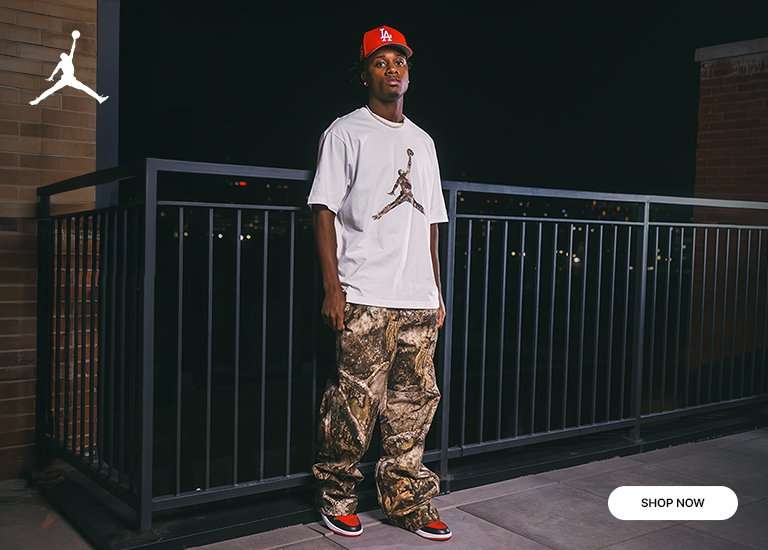 Full-body and close-up view of a person wearing a white t-shirt with a camouflage Jordan logo, camouflage cargo pants, red and black sneakers, and a red baseball cap with a white logo, standing on a rooftop at night.