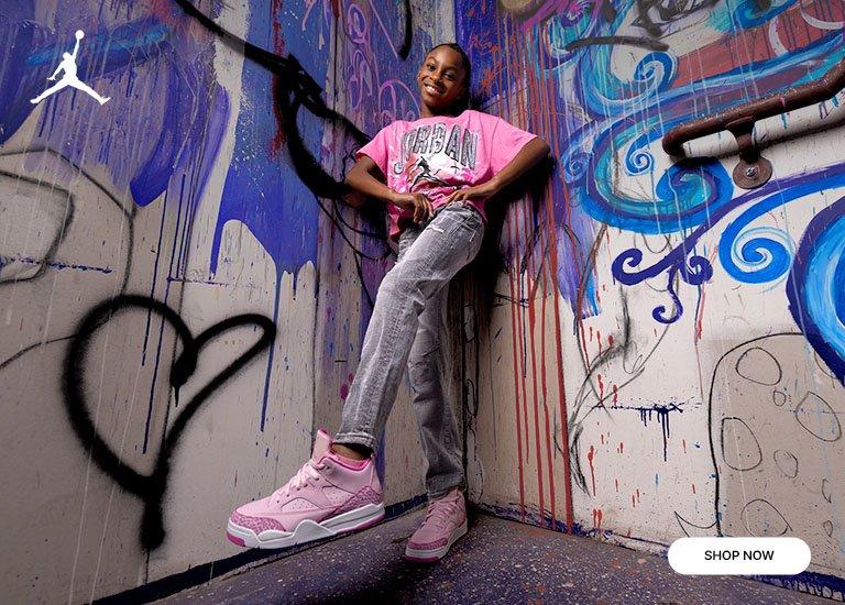 Girl wearing a pink Jordan graphic tee, gray jeans, and pink Jordan sneakers in front of a colorful graffiti wall.