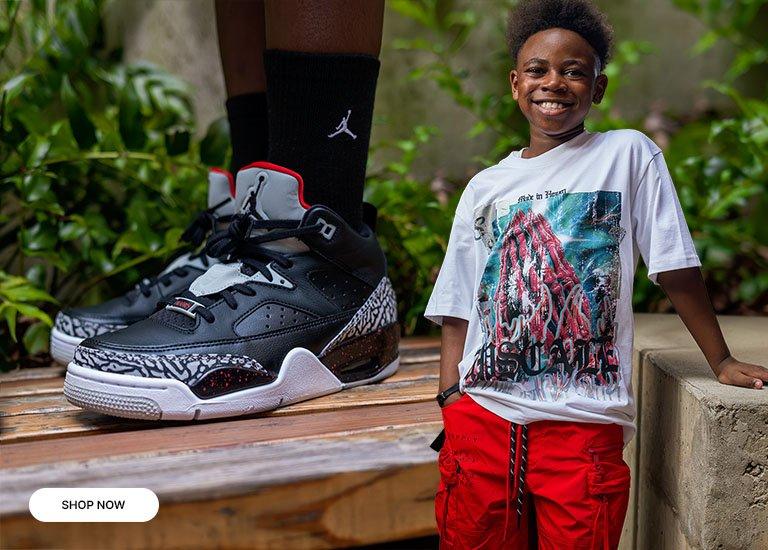 Kid wearing a graphic T-shirt and red cargo shorts stands beside an oversized Jordan Son of Mars sneaker display featuring black, red, and elephant print details in an outdoor setting.