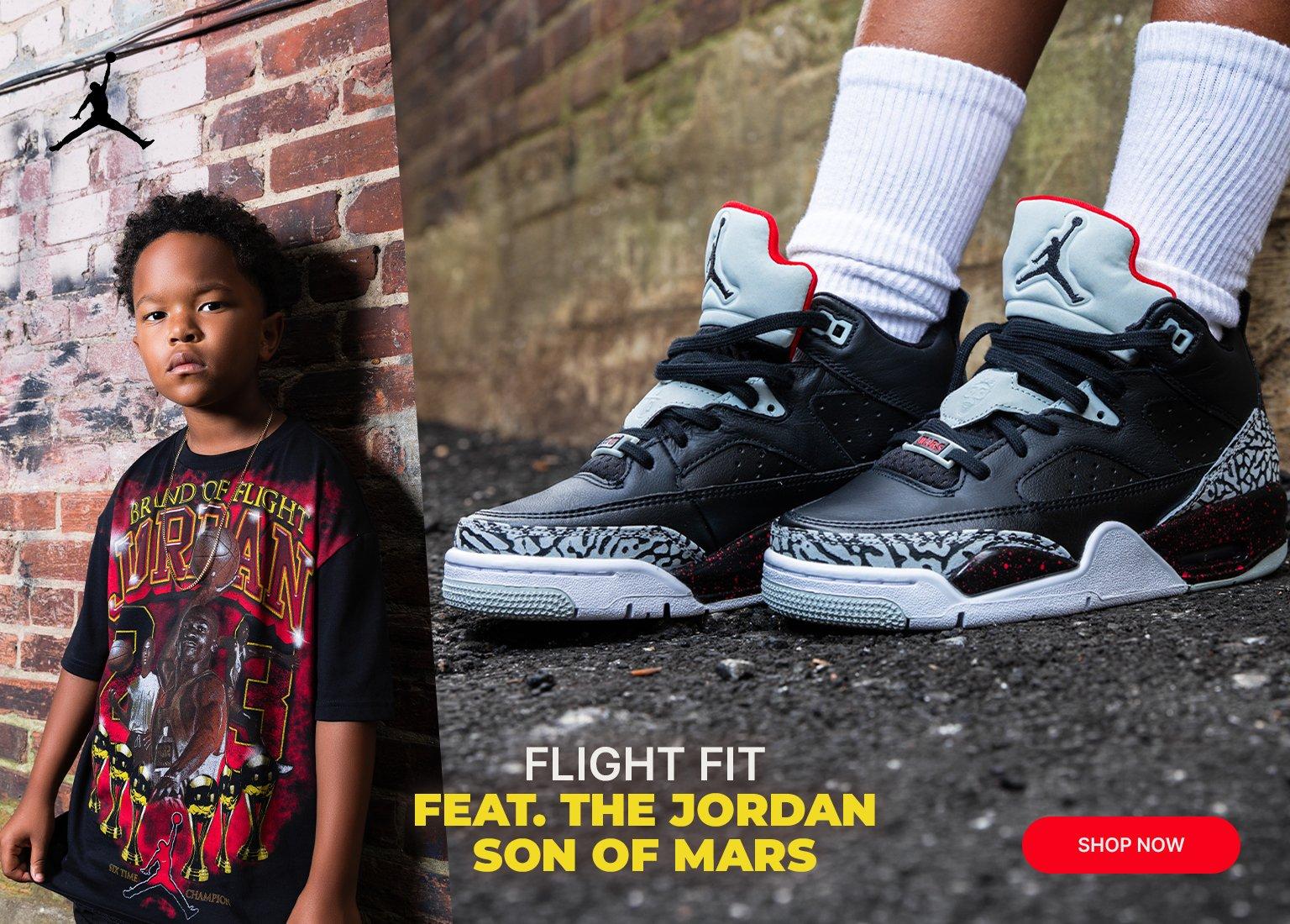 A split-screen promotional banner features a young boy leaning against a brick wall on the left, wearing a black graphic Jordan T-shirt with a black Jordan Jumpman logo in the top corner. On the right, a close-up shows a person wearing the Jordan Son of Mars sneakers in a Black Cement colorway, featuring black leather, grey elephant print overlays, grey tongues with black Jumpman logos, and red accents. Text on the right reads FLIGHT FIT FEAT. THE JORDAN SON OF MARS above a red SHOP NOW button.