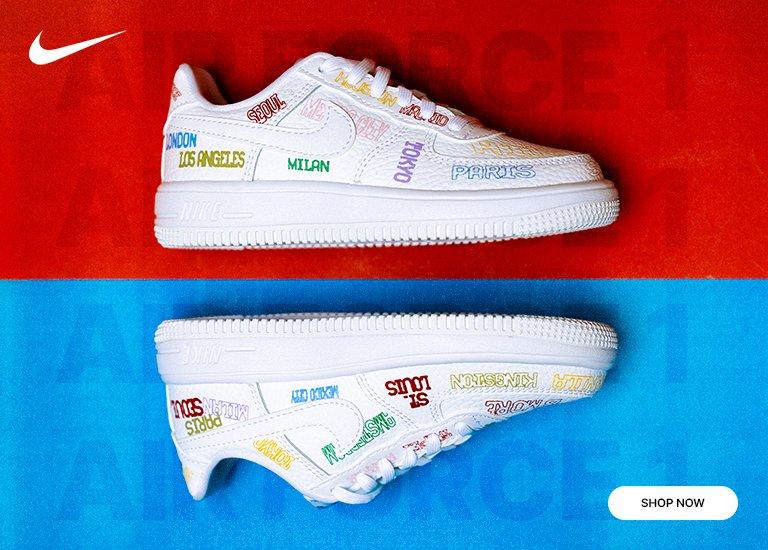 Promotional banner for kids' Nike Air Force 1 "City Pack" sneakers. The image shows two white shoes, one upright on a red background and one inverted on a blue background, both featuring colorful embroidered city names like London, Paris, and Tokyo. Includes a Nike swoosh and a "Shop Now" button.