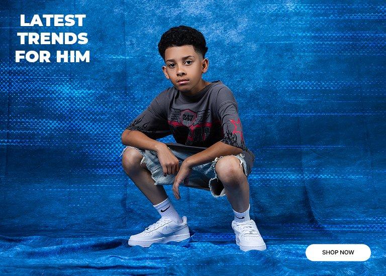 A lifestyle promotional banner for boys' apparel featuring a young boy crouching against a vibrant blue textured background. He is wearing a dark grey graphic tee with red accents, light-wash distressed denim shorts, white Nike crew socks, and white Nike Air Force 1 sneakers. Bold white text in the upper-left corner reads LATEST TRENDS FOR HIM, and a rounded white SHOP NOW button is located in the bottom-right corner.
