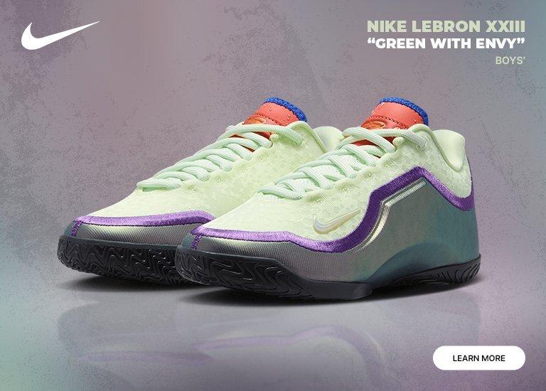  A Nike promotional banner for the Nike LeBron XXIII "Green With Envy" boys' basketball shoes. The image features a pair of low-top sneakers in a pale lime green with iridescent purple and silver accents, orange tongues, and black outsoles. The Nike swoosh and a "LEARN MORE" button are displayed on a textured purple and green background.