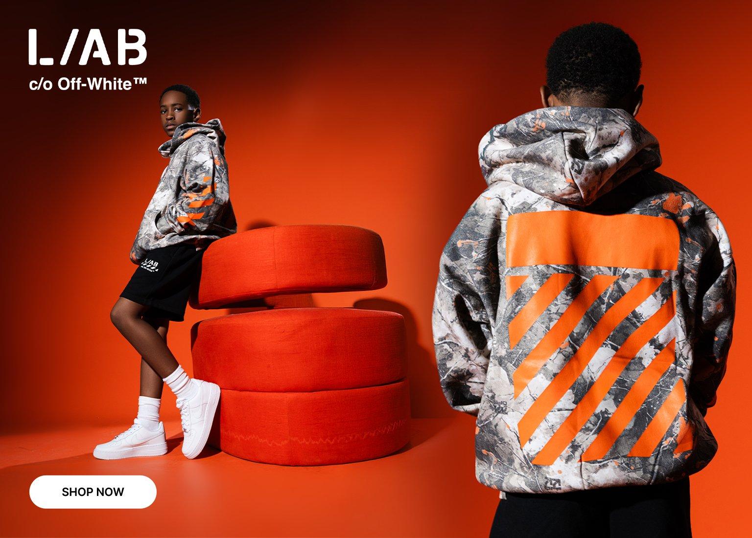 Kids modeling L/A/B c/o Off-White apparel, featuring a gray camo hoodie with bold orange graphics and black shorts against a vibrant orange studio backdrop with modern seating.
