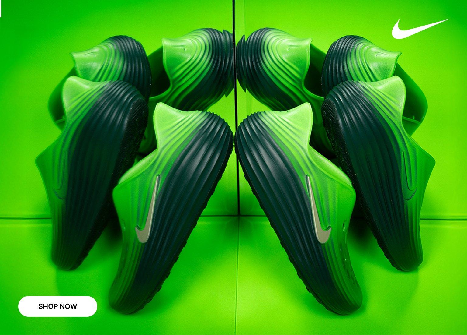 A promotional banner for the kids' Nike Rejuven8 slide, featuring a vibrant green and dark teal colorway. Two pairs of the slip-on shoes are arranged symmetrically on a matching bright green, reflective surface, creating a mirror effect. A white Nike swoosh sits in the top right corner, and a white "SHOP NOW" button is in the bottom left.