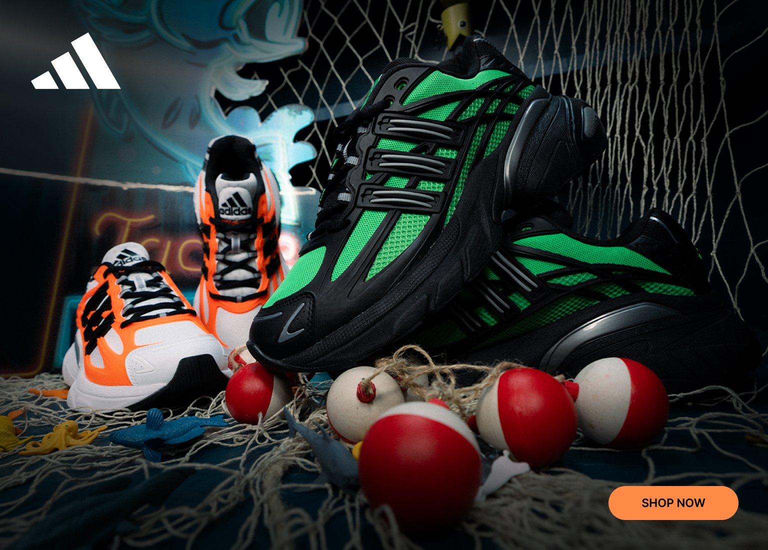 An advertisement for adidas Adistar XLG sneakers featuring two colorways: one in white and orange and the other in black and neon green. The shoes are displayed on a nautical-themed set with fishing nets, bobbers, and small toy fish. The image includes the adidas logo in the top left and an orange "SHOP NOW" button in the bottom right.