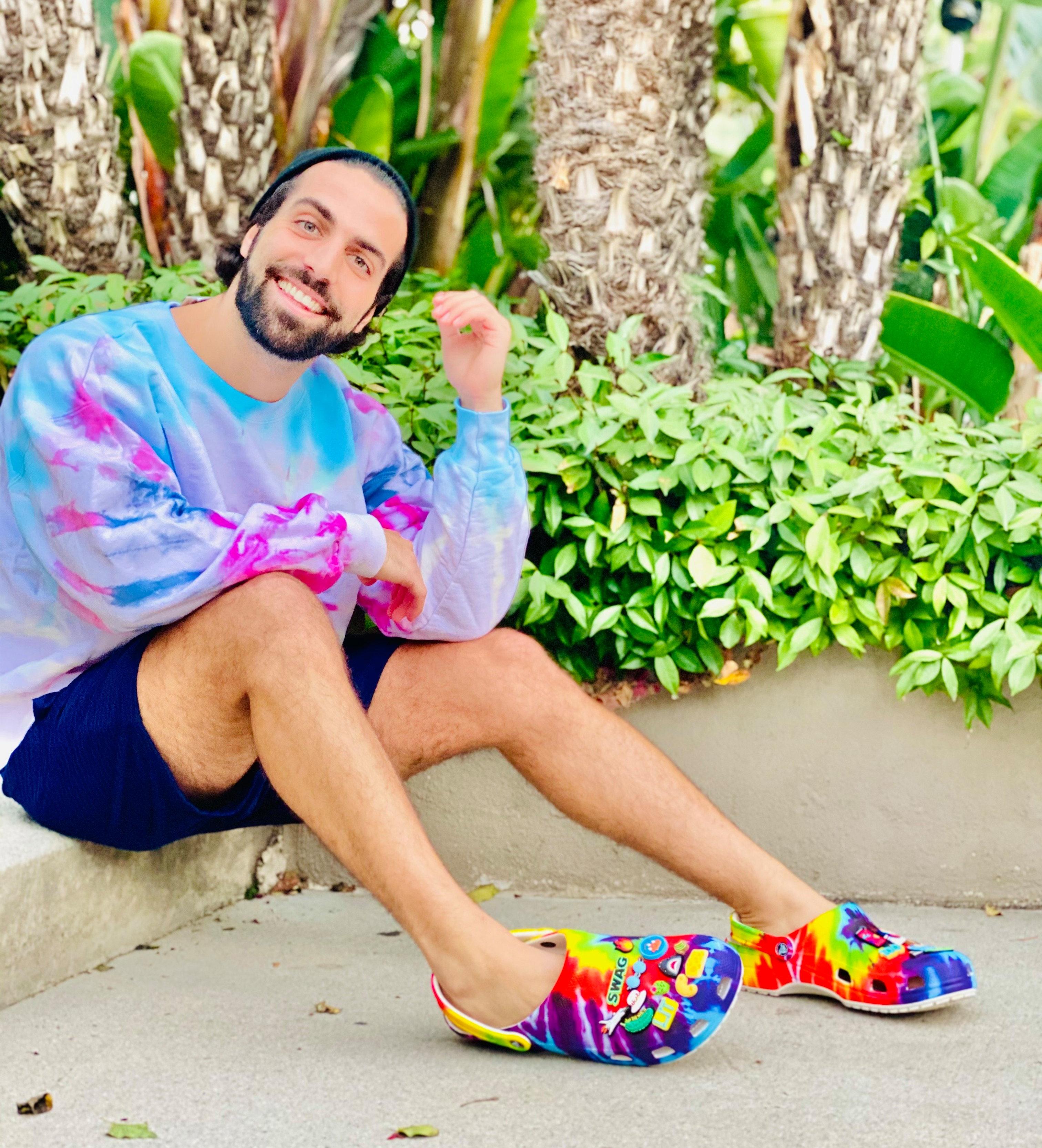 Man sitting on sidewalk in tie-dye sweatshirt, jersey shorts and tie-die Crocs clogs with Jibbitz
