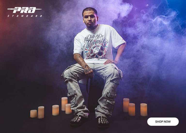 Pro Standard Día de Muertos apparel banner with three models wearing skull-graphic t-shirts and a hoodie against a smoky, dark background with candles. Shop Now button included.