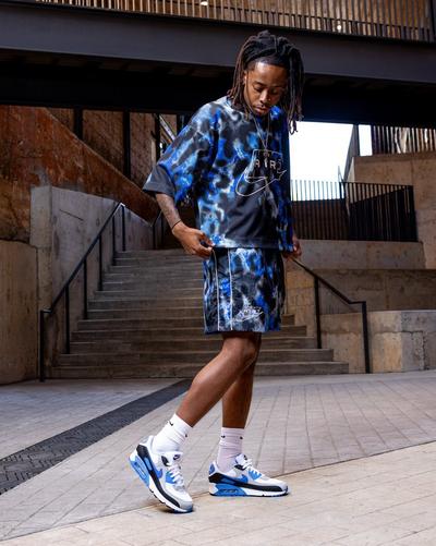 Man standing in an urban stairwell wearing a blue and black Nike matching set with white and blue Nike Air Max sneakers.