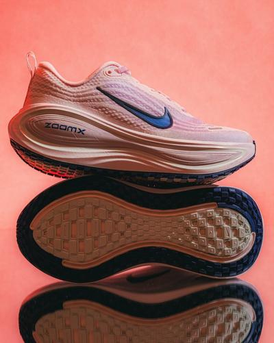 Nike ZoomX running shoe in light pink with a blue Swoosh, shown above a matching outsole on a reflective surface with a pink background.